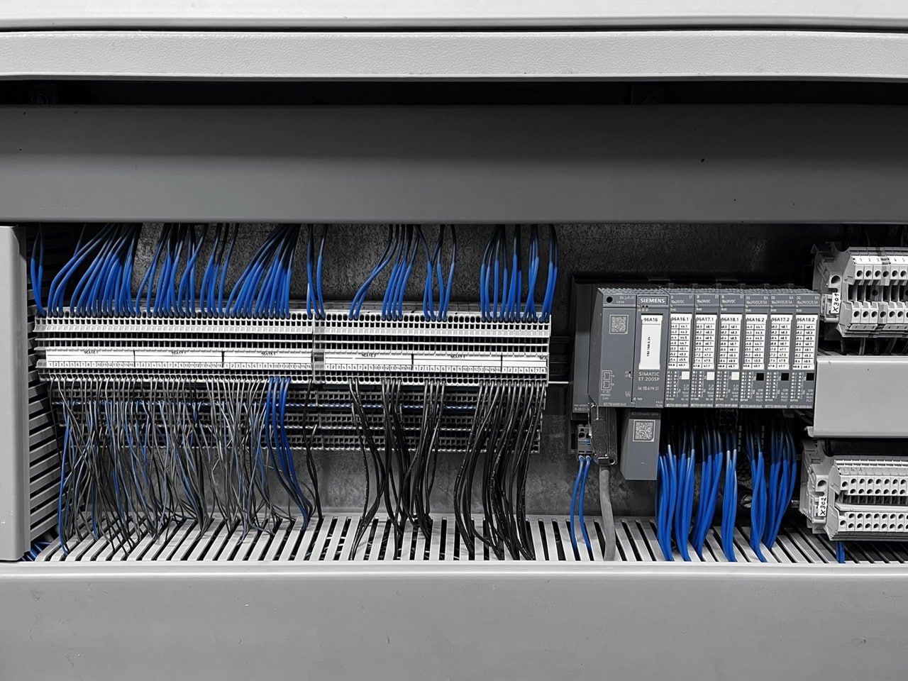 Innenansicht eines Steuerungsschranks mit kabelgebundenen Klemmenleisten, blauen und schwarzen Leitungen und Siemens-SPS-Modulen.
